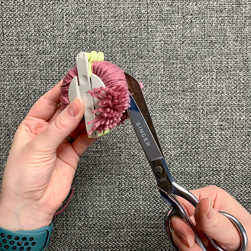 using scissors to cut yarn around the outside of the Pom Pom maker