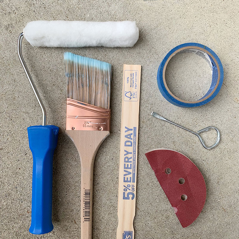 painting supplies on ground: paint brush, roller, can opener, mixing stick, painters tape, and sandpaper