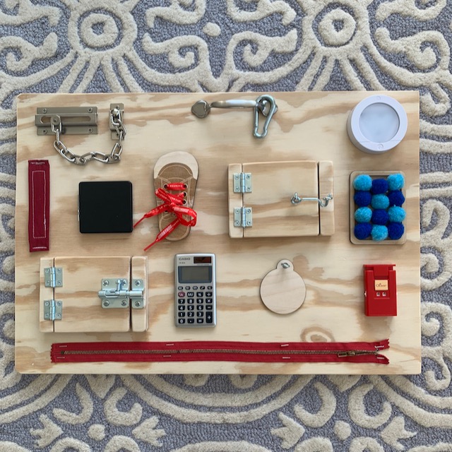 Overhead view of an entire DIY busy board. A piece of wood laid on the floor with several different things for small kids to play with, including: velcro, calculator, knitting stitch counter, zipper, pompoms, puck light, shoe laces, mirror compact, latch pull, carabiner, and latched doors with photos behind.
